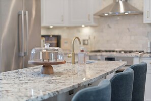 Une cuisine dotée d’un plan de travail en granit, d’un présentoir à gâteaux en bois surmonté d’un couvercle en verre, d’un réfrigérateur en acier inoxydable, d’un robinet doré et d’un flacon de savon liquide.