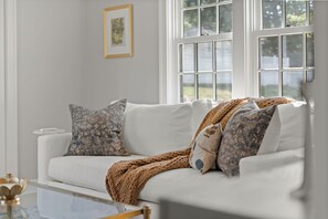 Sofá branco com almofadas estampadas, manta de tricô grosso e um quadro na parede.