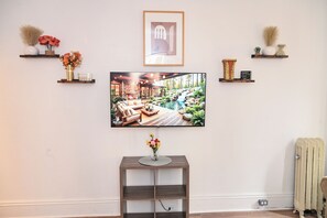 Ein Wohnzimmer mit einem großen Flachbildfernseher an der Wand, einem hölzernen Couchtisch mit Blumen in einer Vase und einer Heizung am Boden.
