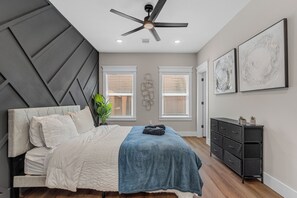 Ein Schlafzimmer mit einem Bett, einem Deckenventilator, zwei Fenstern mit Jalousien, einem Schminktisch und Wandbildern.