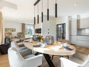 A modern dining area with a round table, multiple chairs, pendant lights, and a kitchen in the background. - Holiday Home Sylt 44 (Wenningstedt-Braderup)