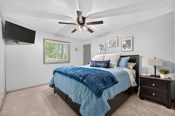 A bedroom with a bed, a nightstand, a lamp, a ceiling fan, a TV, and artwork on the wall.