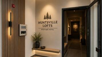 A hotel lobby with a reception desk, a wall-mounted fire alarm, a potted plant, and a sign for Huntsville Lofts Boutique Hotel.