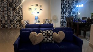 Un salón con sofá azul, cojines con forma de corazón, televisor montado en la pared y cortinas estampadas.