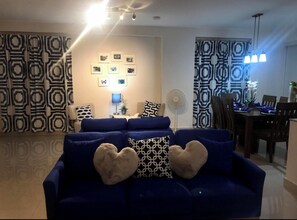 Uma sala de estar com um sofá azul, almofadas em formato de coração, televisão na parede e cortinas estampadas.