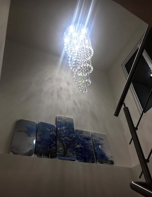 A chandelier with multiple light fixtures hanging from the ceiling, a set of three framed artworks on the wall below, and a window to the right.