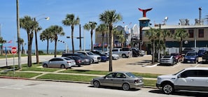 A parking lot with multiple cars, palm trees, and a building with a distinctive red tail fin. - Pleasure Pier View 2BR · Steps to Seawall (Galveston)