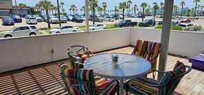 A covered outdoor seating area with a round table and striped chairs, overlooking a parking lot with multiple cars and palm trees. - Pleasure Pier View 2BR · Steps to Seawall (Galveston)