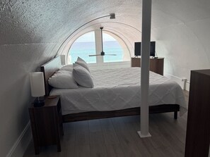 A bedroom with a bed, a nightstand, a lamp, a view of the sea through a circular window, and a wooden floor. - Newly Remodeled & Pet Friendly - Sunset Surfside (Panama City Beach)