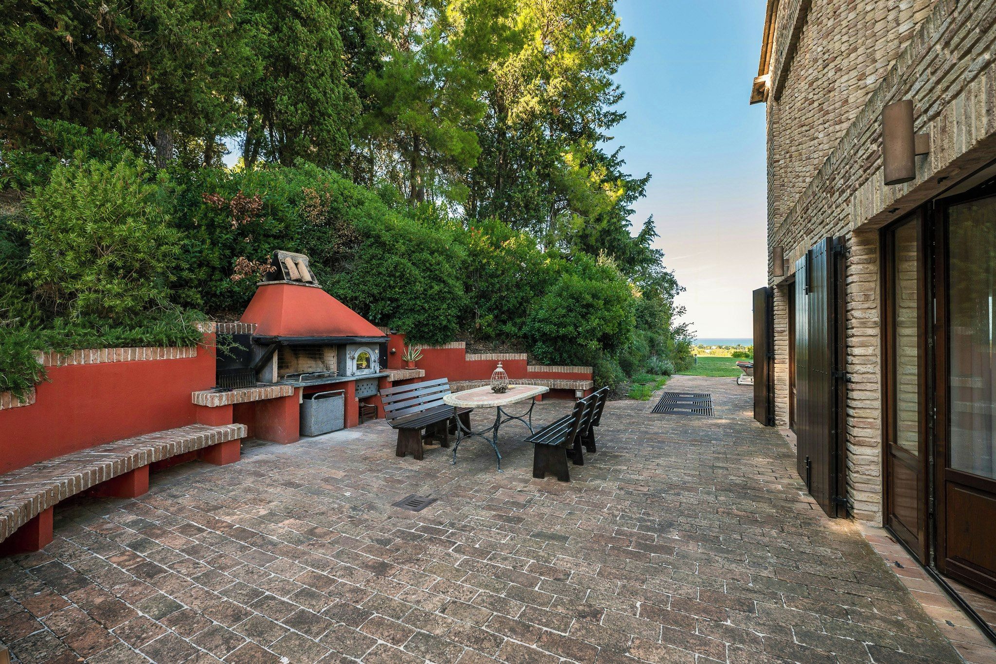 Een terras buiten met een betegelde vloer, een barbecue en een eettafel met banken.