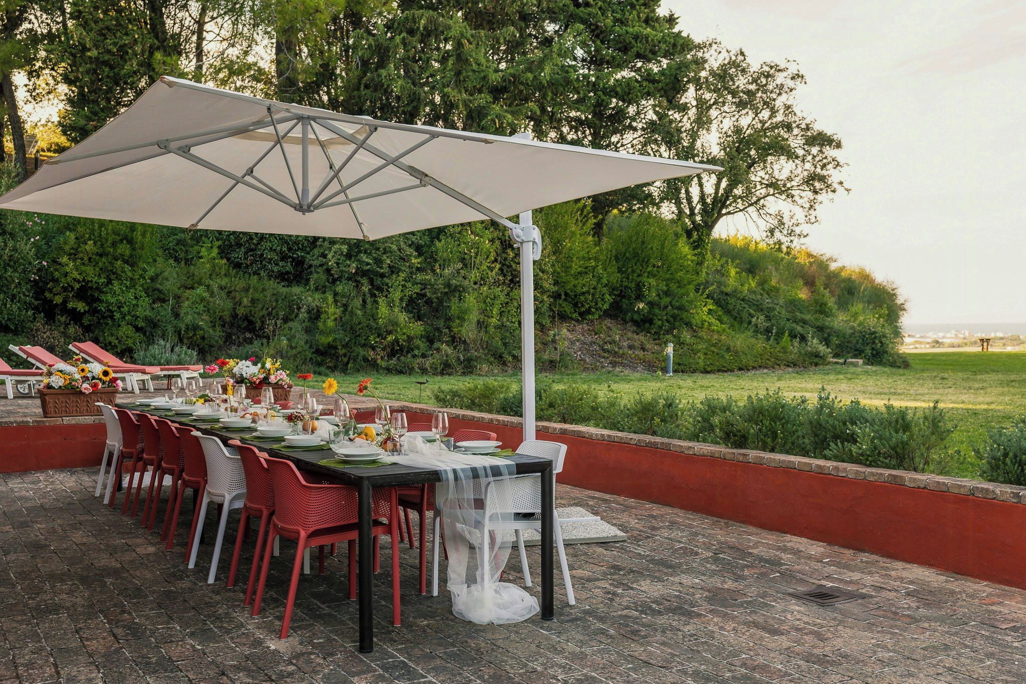 Un espace de restauration en plein air avec une longue table dressée pour un repas, entourée de chaises, et un grand parasol offrant de l’ombre.