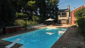 Une piscine rectangulaire bordée de briques, entourée de chaises longues et d’une table placée sous une pergola.
