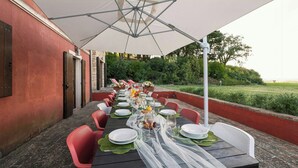 Ein Essbereich im Freien mit einem langen Tisch, gedeckt für eine Mahlzeit, umgeben von Stühlen und einem Sonnenschirm, der Schatten spendet.