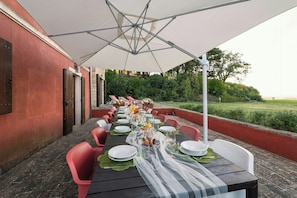 Ein Essbereich im Freien mit einem langen Tisch, gedeckt für eine Mahlzeit, umgeben von Stühlen und einem Sonnenschirm, der Schatten spendet.