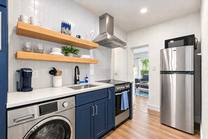 A modern kitchen with a stainless steel refrigerator, a built-in oven, a washing machine, and wooden shelves above the counter. - Steps to St. Pete Beach / Luxury Coastal Condo #2 (St. Pete Beach)