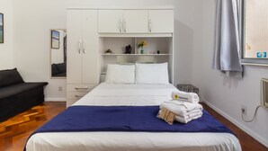 Una habitación de hotel con cama, armario, espejo, ventana con cortinas y una mesita con un jarrón de flores.