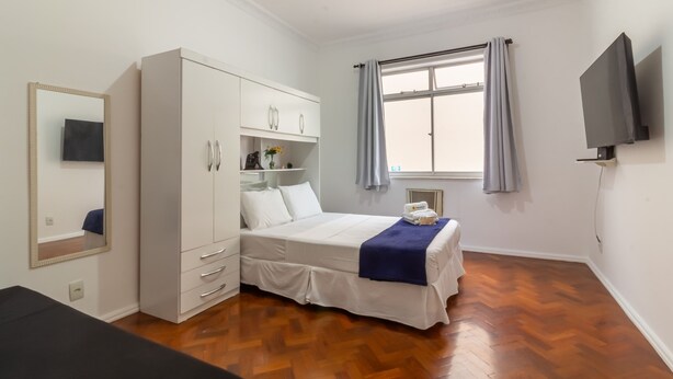 Un dormitorio con cama, armario, espejo, televisor y ventana con cortinas.