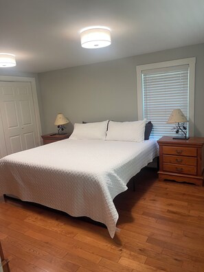 A bedroom with a bed, two bedside tables, a window with blinds, and a door. - OVERSIZED 1BR/1BA Winter Seasonal Rental (New Hampton)