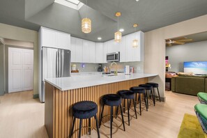 A modern kitchen with white cabinets, a stainless steel refrigerator, a microwave, and a bar area with stools. - Heated Pool + Spa | Mini Golf | 5BR Coastal Retreat Near Anna Maria Island (Bradenton)