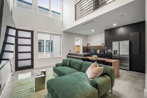 Un salon moderne avec un canapé vert, une cuisine aux meubles sombres et une vue sur le ciel par de grandes fenêtres.