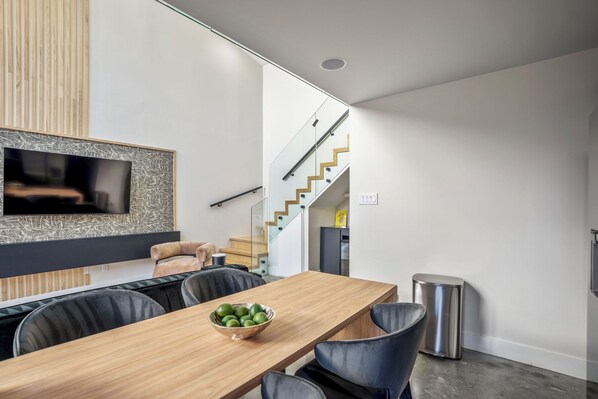 A modern dining area with a wooden table, black chairs, and a bowl of fruit. A staircase with glass railings leads to an upper level.