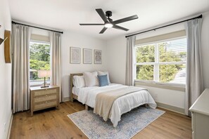 Une chambre à coucher comprenant un lit, une table de chevet, une commode, un ventilateur de plafond, une fenêtre avec stores et des œuvres d’art murales.