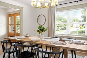 A dining room with a long table set for six, surrounded by black chairs, a chandelier, and a potted plant.