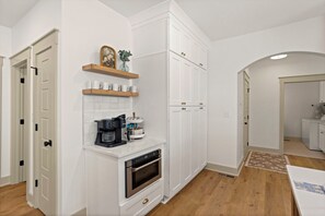 Une cuisine moderne avec des meubles blancs, un four encastré, une machine à café et une petite étagère avec des tasses.