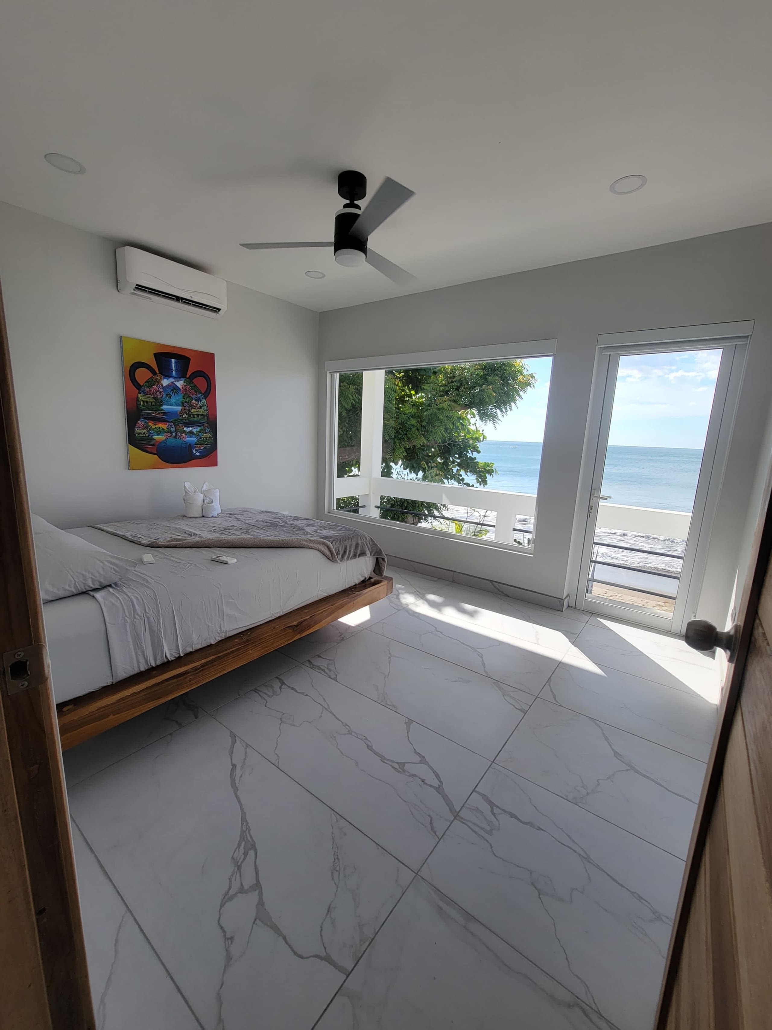A bedroom with a bed, a ceiling fan, a painting, and a view of the sea.