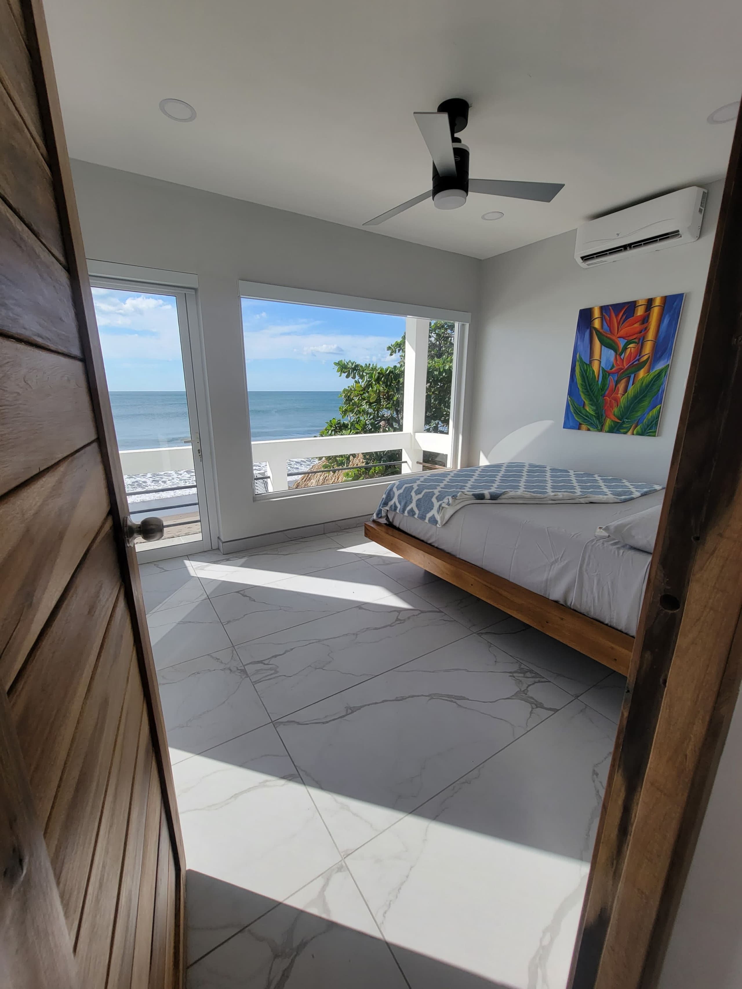 Een slaapkamer met een bed, een plafondventilator, een schilderij en uitzicht op de oceaan.