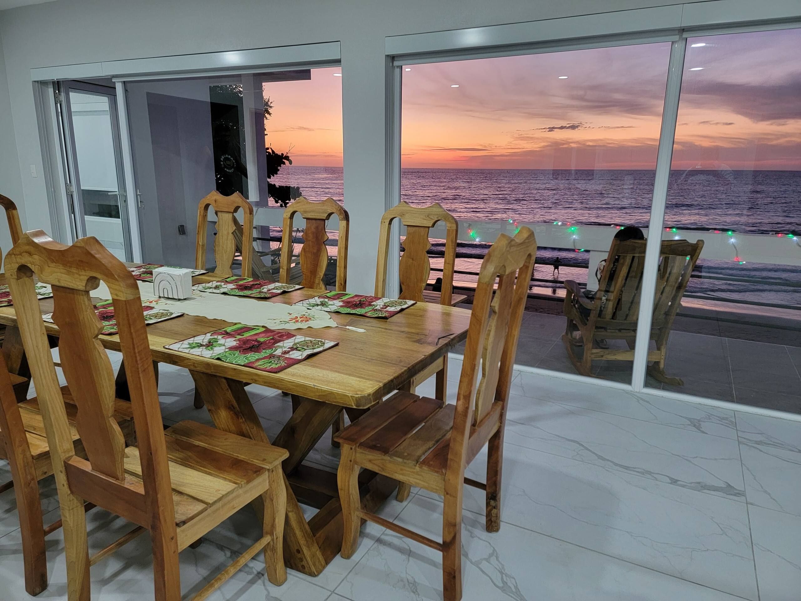 Un coin repas avec vue sur la mer au coucher du soleil, des chaises en bois, une table dressée avec des sets de table et une porte vitrée donnant sur l’extérieur.