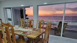 A dining area with a view of the sea at sunset, wooden chairs, a table set with placemats, and a glass door leading outside. - 7-bedroom Beach Front house in Play Miramar with AC (Miramar)