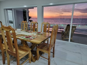Comedor con vista al mar al atardecer, sillas de madera, una mesa con individuales y una puerta de vidrio que da al exterior.
