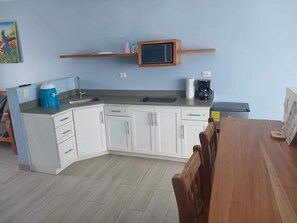 Une cuisine compacte avec des meubles blancs, un plan de travail en bois, une machine à café et un petit téléviseur posé sur une étagère.
