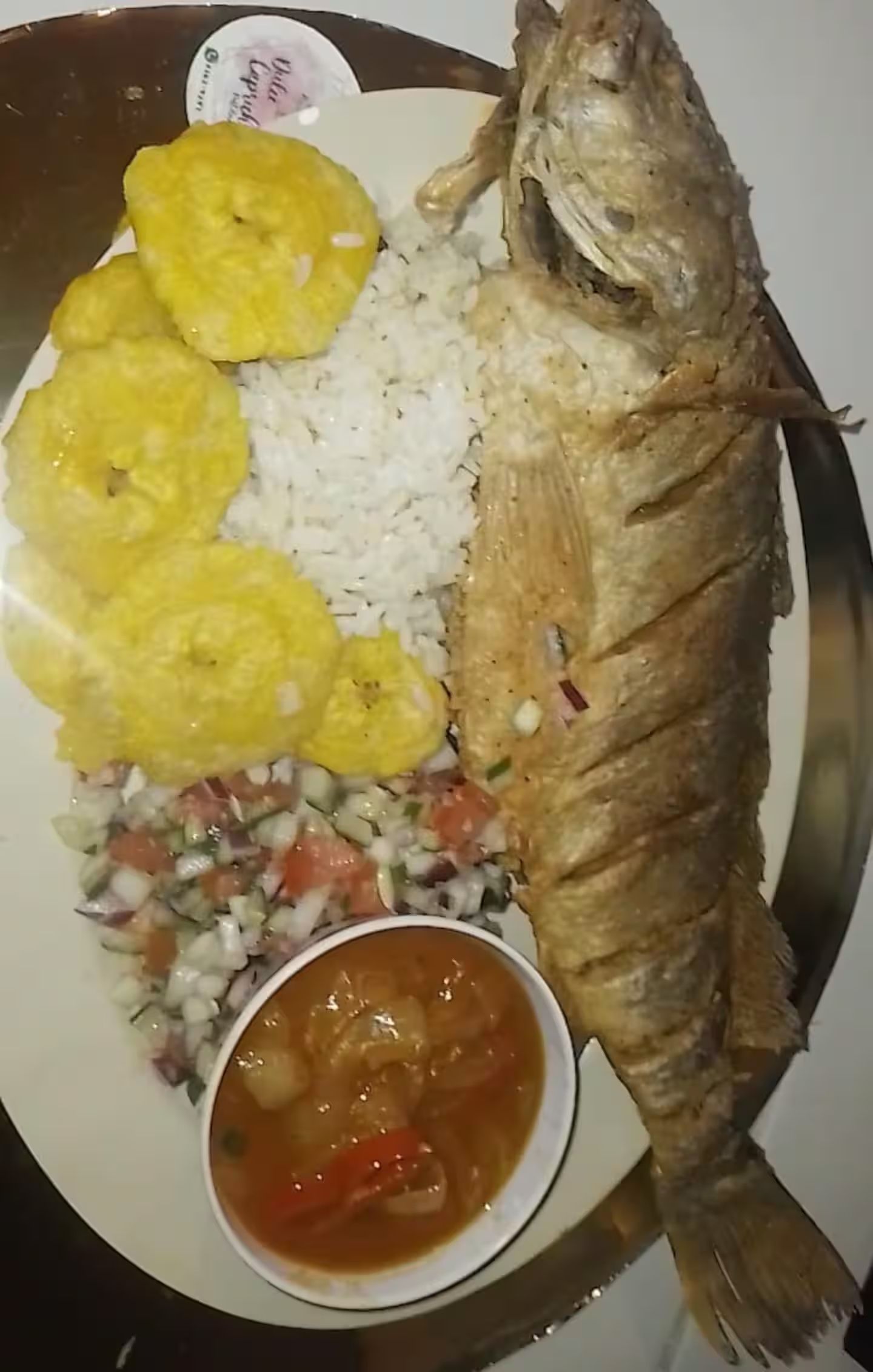 En tallrik med en hel, friterad fisk, ris, friterade bananer, en sallad och en liten skål med sås.