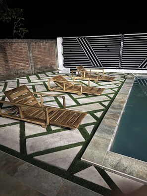 A poolside area with a geometric patterned patio, wooden lounge chairs, and a brick wall.