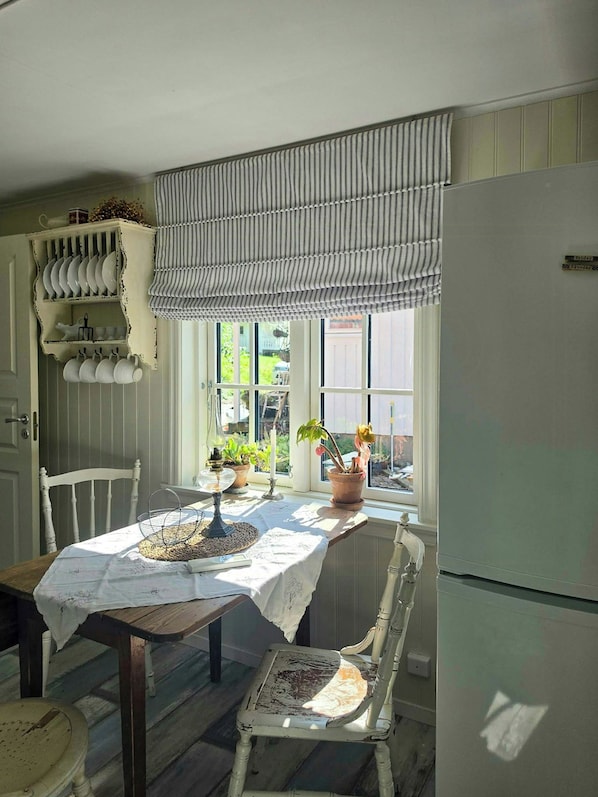 A dining area with a table set for two, a window with a view of greenery, and a white refrigerator. - Renovated guesthouse with courtyard in Ljungskile (Västra Götalands län)