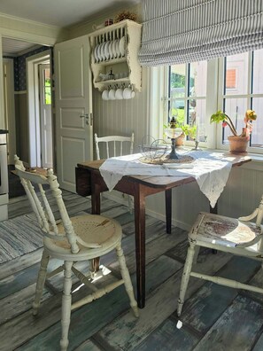 A dining area with a table set for two, surrounded by vintage chairs, a window with a view, and a shelf with dishes. - Renovated guesthouse with courtyard in Ljungskile (Västra Götalands län)
