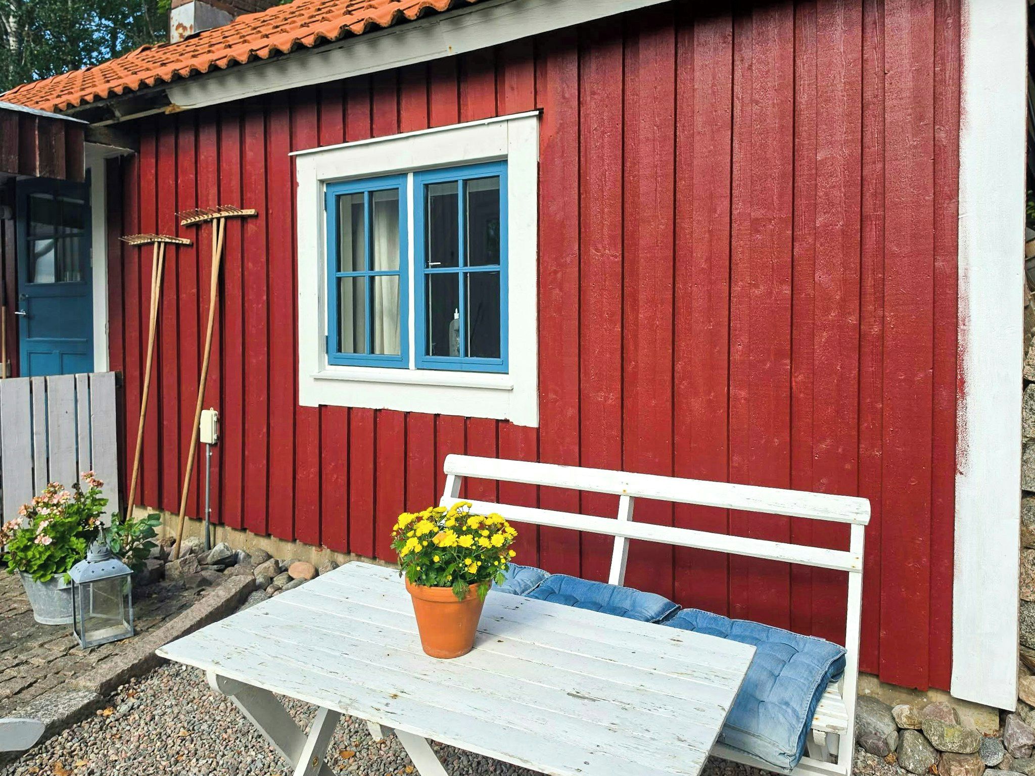 Een rood houten gebouw met een witte bank, een tafel, een blauw raam en een bloempot met gele bloemen.