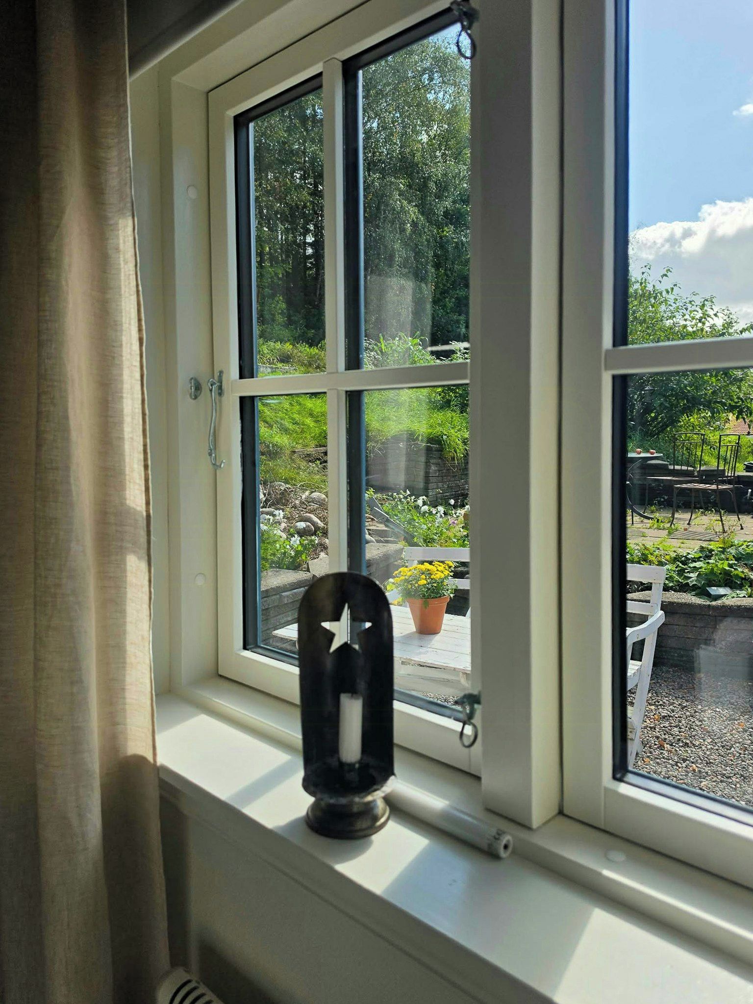 Ett fönster med utsikt över en trädgård, en vit stol och ett svart föremål på fönsterbrädan.