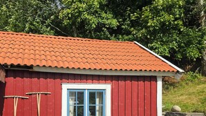 Ein rotes Holzgebäude mit Ziegeldach, einem Fenster mit blauer Umrandung, einer weißen Bank und einem Tisch mit einem Blumentopf.