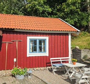 A red wooden building with a tiled roof, a window with blue trim, a white bench, and a table with a potted plant. - Renovated guesthouse with courtyard in Ljungskile (Västra Götalands län)