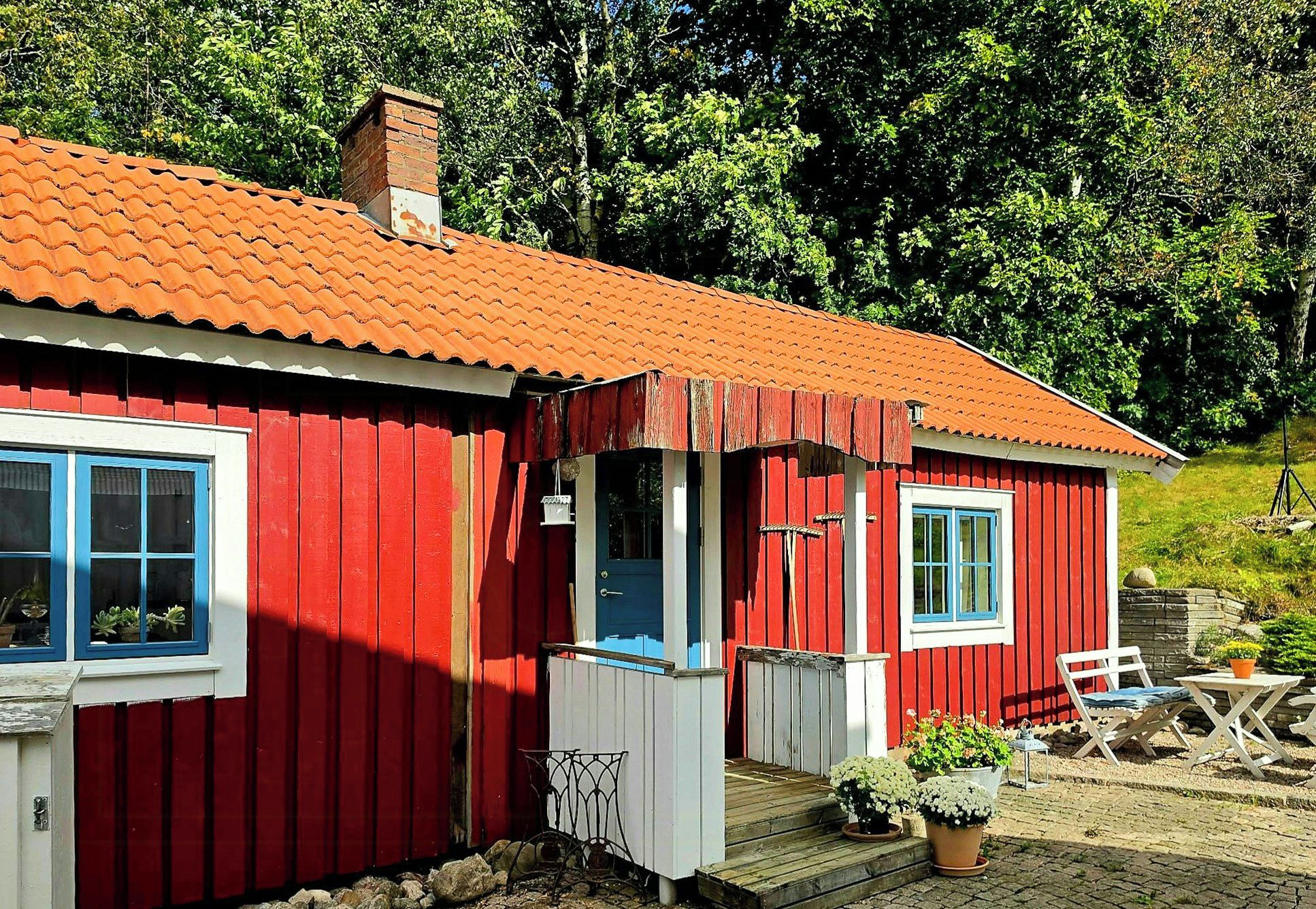 A red wooden house with a tiled roof, a porch, and a small garden area with a table and chairs.