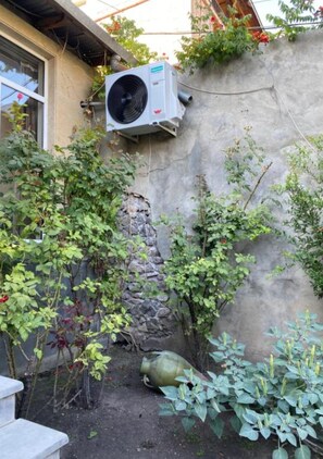 A garden area with a stone wall, a large air conditioning unit mounted on the wall, and various plants including a flowering bush and a large green plant with yellow flowers. - Alikhanyan House (Yerevan)