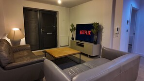 Sala de estar com sofá, mesa de centro, TV exibindo 'NETFLIX' e luminária.