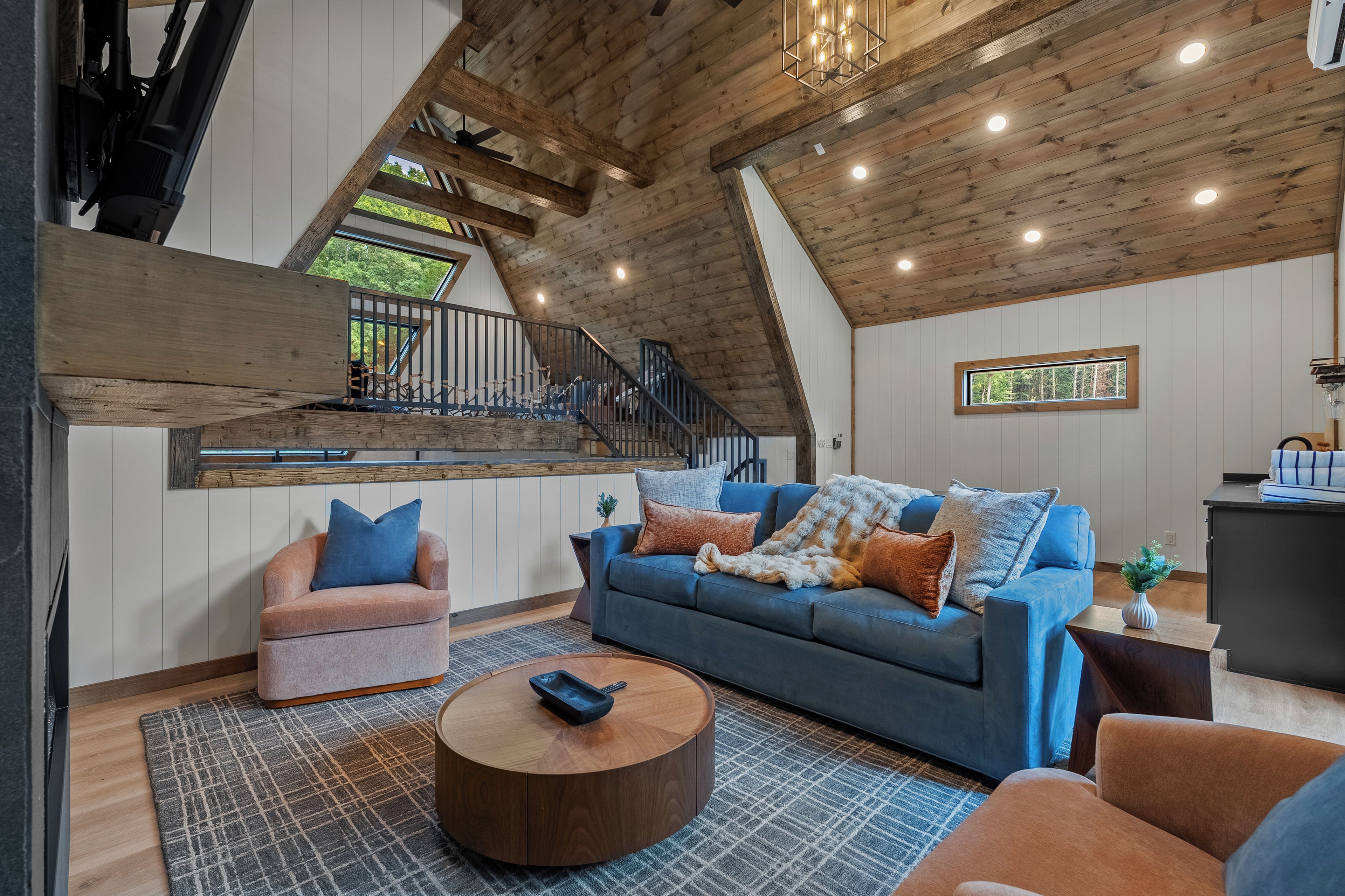 A loft-style living room with a high ceiling, wooden beams, a blue sofa, a pink armchair, and a round coffee table.