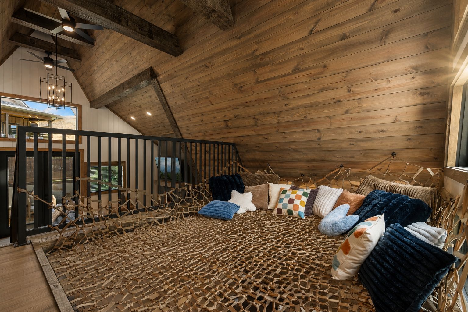 Un dormitorio acogedor en el ático, con techo inclinado, vigas de madera, una cama con varios cojines y vistas al exterior a través de una ventana.