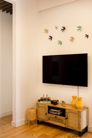 Un salon doté d’un téléviseur à écran plat fixé au mur, d’une console en bois agrémentée d’objets décoratifs et d’un petit fauteuil en osier.