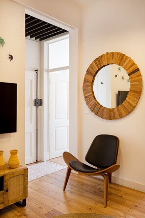 Une pièce avec un plancher en bois, une chaise noire, un miroir rond, un téléviseur et une porte donnant sur une autre pièce.