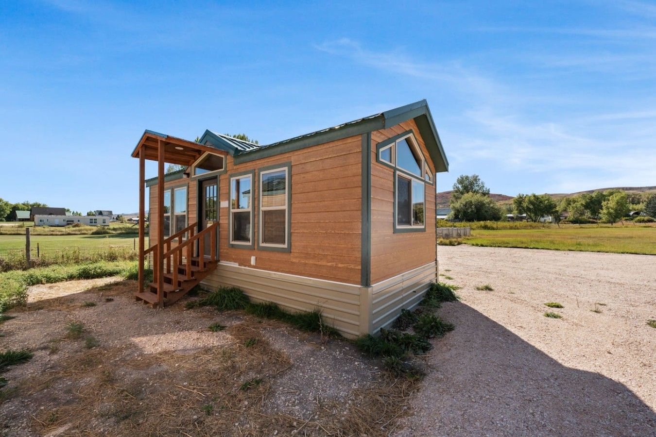 Una pequeña cabaña de madera con un porche y una escalera, ubicada en una zona rural con campos abiertos.
